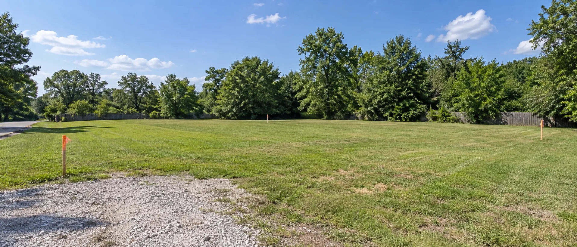 A mowed vacant lot marked with survey stakes, bordered by mature trees
